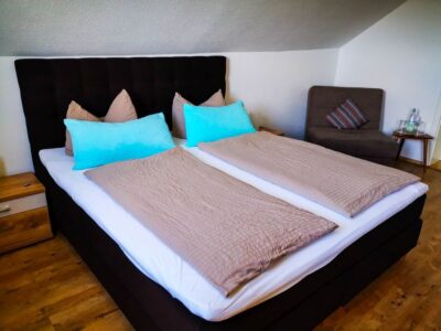 Ein ordentlich gemachtes Doppelbett mit türkisfarbenen Kissen und beigen Bettdecken steht in einem gemütlichen Erfurter Zimmer mit Holzfußboden, einem grauen Sessel mit gestreiftem Kissen und Nachttischen auf jeder Seite.