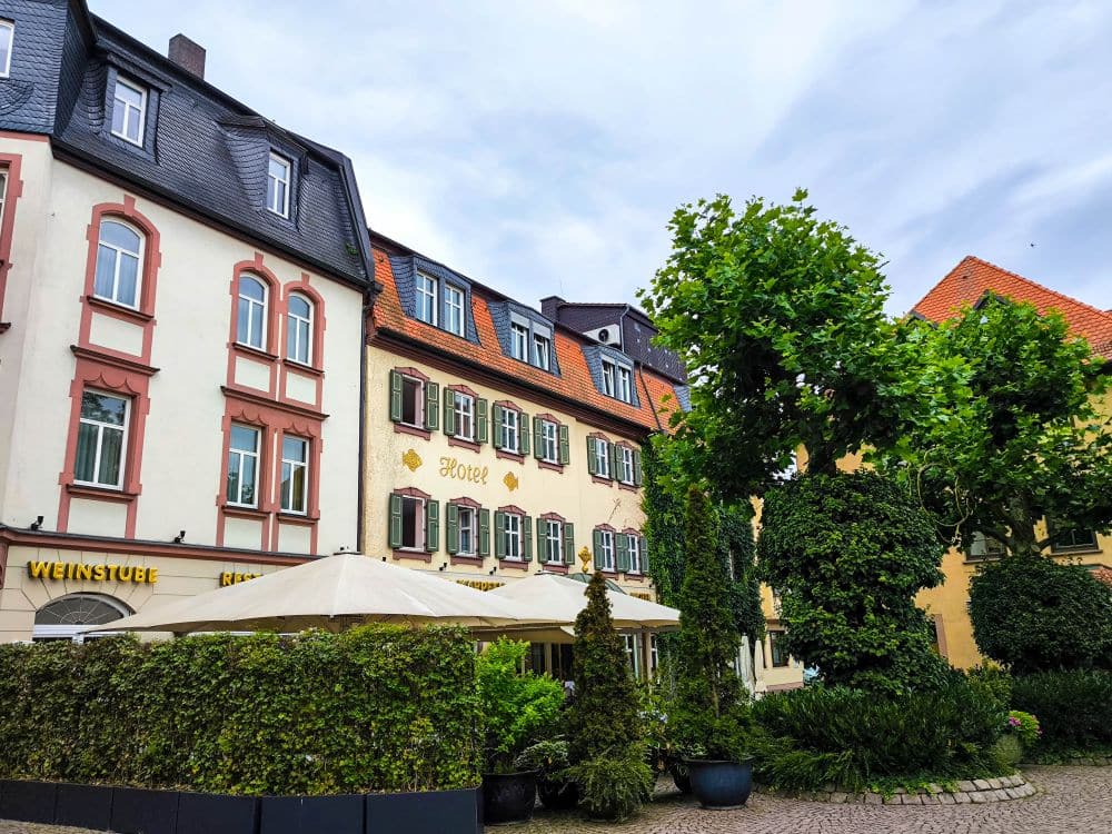 Eine charmante Straßenszene in Fulda mit historischen Gebäuden, einem Hotel, grünen Bäumen und einer von großen Sonnenschirmen beschatteten Terrasse eines Restaurants. Der Himmel ist teilweise bewölkt und schafft eine friedliche und einladende Atmosphäre.