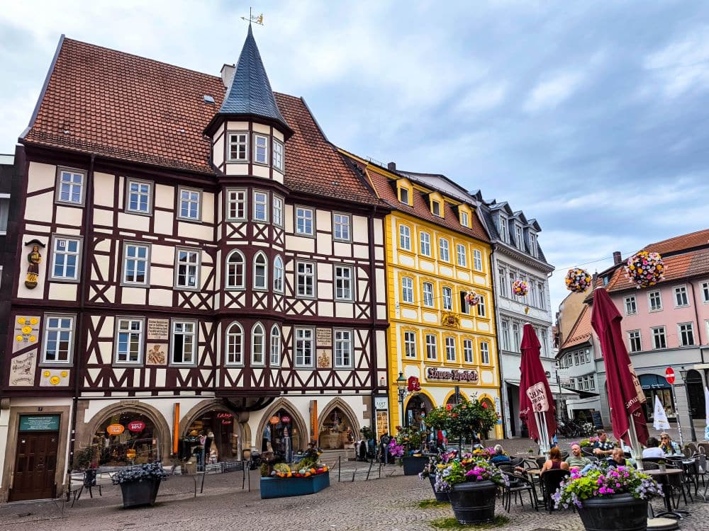 Ein lebendiger europäischer Platz in Fulda mit Fachwerk und farbenfrohen historischen Gebäuden, Cafétischen im Freien, speisenden Menschen, Blumenkübeln und einem wolkenverhangenen Himmel über uns.