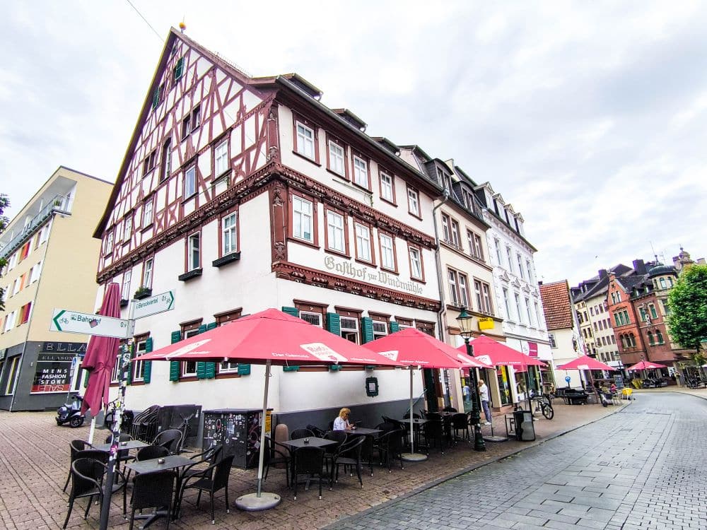 Ein Fachwerkhaus mit Sitzgelegenheiten im Freien und roten Sonnenschirmen steht an einer Straßenecke in Fulda, wo die Menschen an Tischen neben den bunten Gebäuden sitzen, die die kopfsteingepflasterte Straße säumen.