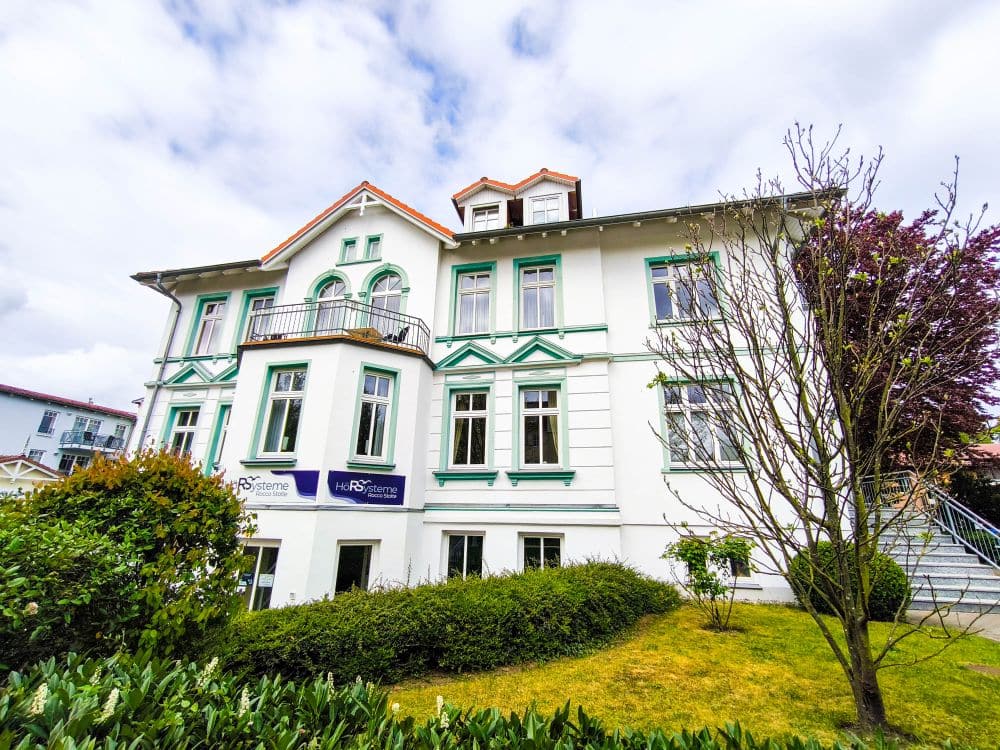 Ein großes, weißes, zweistöckiges Gebäude mit grüner Verkleidung, hohen Fenstern, einem kleinen Balkon und gepflegten Sträuchern steht vor dem teilweise bewölkten Himmel in Kühlungsborn - "Hansezimmer" steht auf den Schildern und lädt zu einer Auszeit an der Ostsee ein.