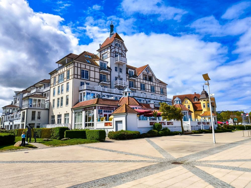 Unter einem teilweise wolkenverhangenen blauen Himmel steht in Kühlungsborn an der Ostsee ein prächtiges, weißes Hotel am Meer mit Fachwerkakzenten und Balkonen. Das Gebäude ist von Grünflächen und einem breiten gepflasterten Weg umgeben - perfekt für eine entspannte Auszeit.