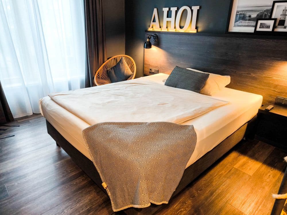 Ein modernes Hotelzimmer in Ludwigsburg mit einem sauber gemachten Doppelbett, grauer und weißer Bettwäsche, einem dekorativen Kissen und einem Korbsessel. Beleuchtete "AHOI"-Schriftzüge auf einer Akzentwand aus dunklem Holz erinnern an die Eleganz des Barock. Ein großes Fenster mit transparenten Vorhängen lässt natürliches Licht herein.