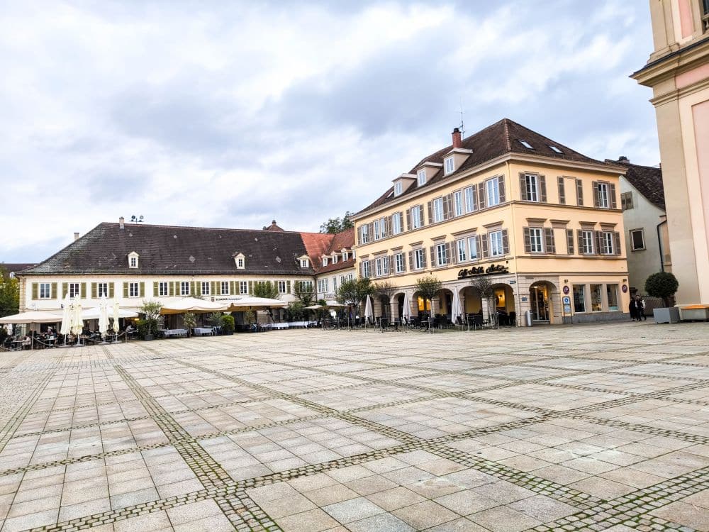 Ein großer, leerer Platz mit Kopfsteinpflaster in Ludwigsburg, gesäumt von europäischen Gebäuden aus der Barockzeit mit Cafés und Sitzgelegenheiten im Freien unter einem bewölkten Himmel, erinnert an den Charme der historischen Gärten.