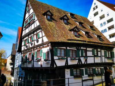 Ein historisches Fachwerkhaus mit steilem Ziegeldach und grünen Fensterläden steht neben modernen weißen Gebäuden unter einem blauen Ulmer Himmel; im Vordergrund sind Menschen zu sehen.