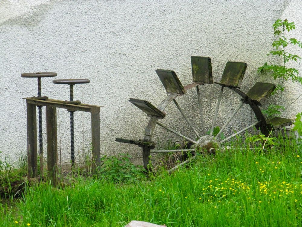 Ein altes, verwittertes Wasserrad und rostige Metallmaschinen stehen im überwucherten Gras neben einer strukturierten weißen Wand und bieten einen Einblick in die Geschichte Ihrer Kleinen Lautertal Wanderung - der perfekte Ort für eine ruhige Auszeit inmitten von Grünpflanzen.