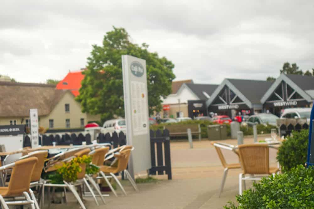 Außensitzplatz eines Cafés mit leeren Tischen und Stühlen, Grünzeug und einer Menütafel im Vordergrund. Im Hintergrund sind Geschäfte mit dem Namen WESTWIND und geparkte Autos zu sehen - sie fangen den Charme von Blavand in Dänemarks wildromantischem Westen ein.