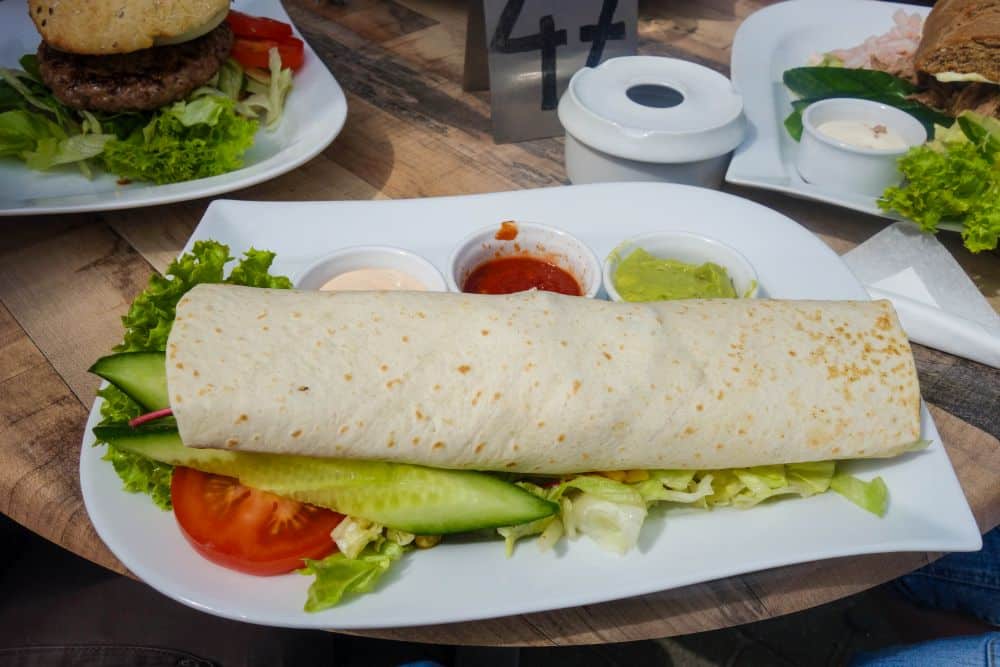 Ein großer Tortilla-Wrap, gefüllt mit frischem Salat, Gurke und Tomate, wird auf einem weißen Teller mit drei kleinen Behältern mit Soßen und Dips serviert und erinnert an den wildromantischen Charme von Dänemarks Westen. Ein weiteres Gericht ist teilweise im Hintergrund zu sehen.