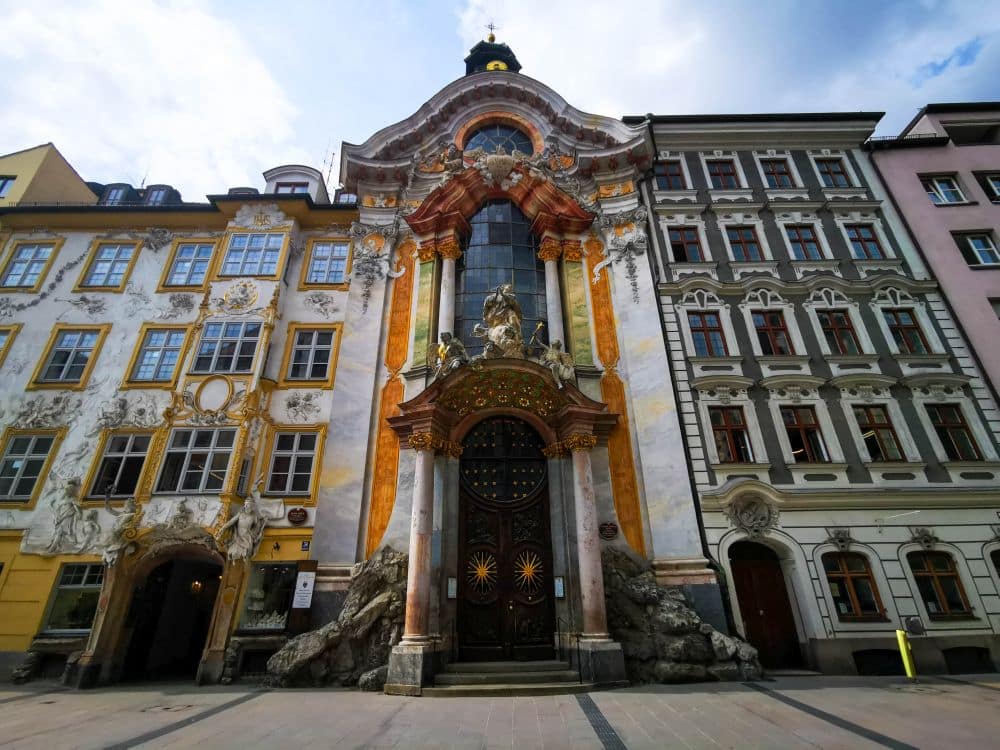 Barocke Kirche in München mit verzierter Fassade, gewölbten Holztüren, goldenen und orangefarbenen Verzierungen, flankiert von dekorativen historischen Gebäuden unter einem teilweise bewölkten Himmel. Stufen und felsähnliche Elemente führen zum Eingang.