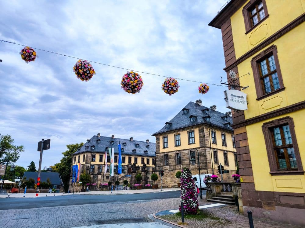 Eine europäische Straßenszene in Fulda mit historischen gelben Gebäuden, einer blumengeschmückten Statue und bunten kugelförmigen Blumenarrangements, die unter einem bewölkten Himmel über der Straße hängen.