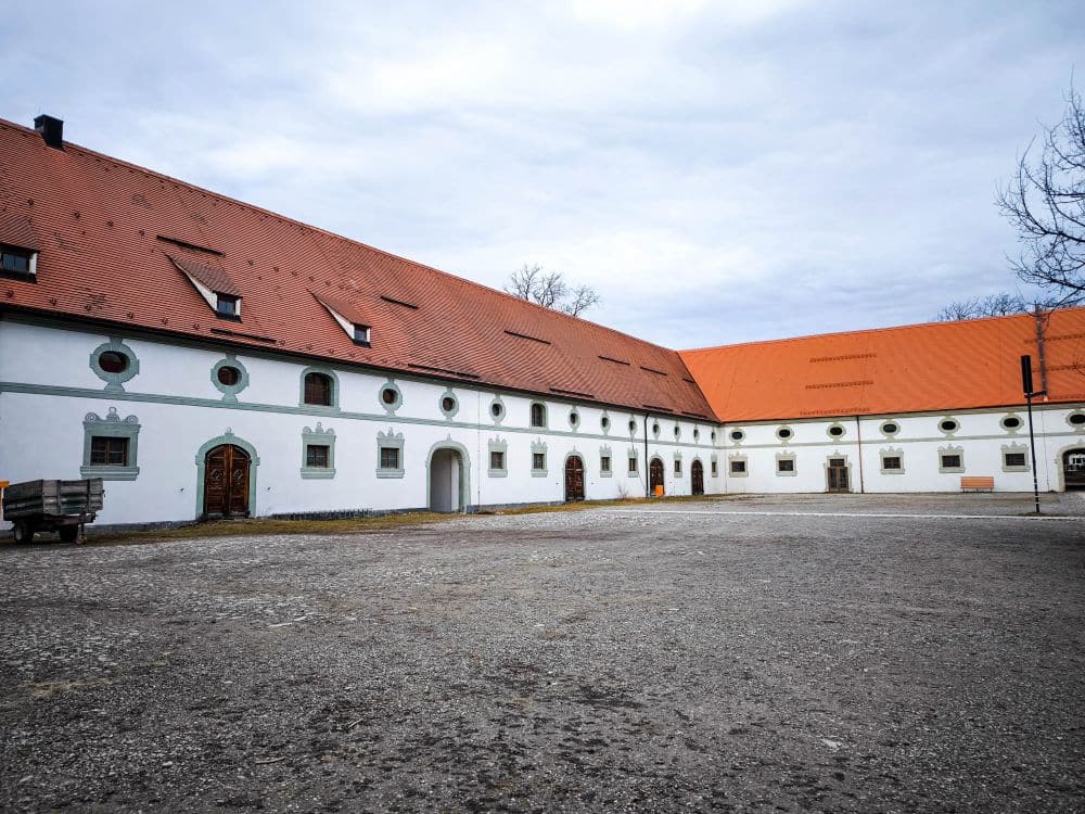 Ein großes Barockgebäude mit weißen Wänden, grüner Verkleidung und rotem Ziegeldach - ein Teil des Klosters Benediktbeuern - umgibt einen geräumigen Kieshof unter einem bewölkten Himmel. An der Fassade sind mehrere Holztüren und kleine Fenster zu sehen.