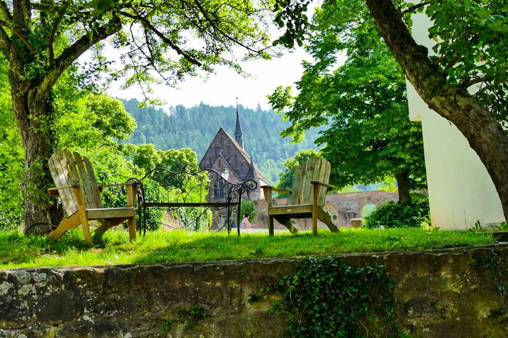 Zwei Holzstühle und eine Metallbank stehen auf einer von Bäumen beschatteten Wiesenterrasse mit Blick auf die kleine Steinkirche mit Kirchturm in Hirsau, umgeben von üppigem Grün und bewaldeten Hügeln.