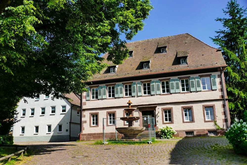 Ein historisches Hirsauer Gebäude mit braunen Dachziegeln, grünen Fensterläden und weißen Wänden steht hinter einem steinernen Brunnen, der teilweise eingezäunt ist; ein großer Baum spendet Schatten unter dem klaren blauen Himmel.