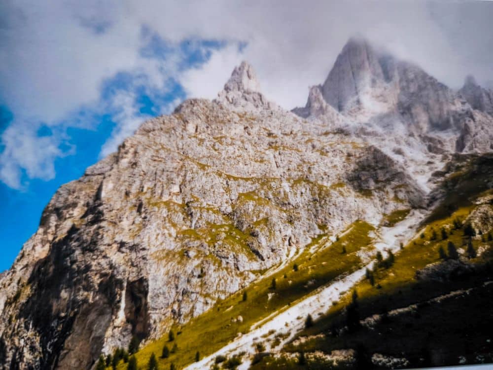 Ein felsiger Berggipfel, der teilweise von Wolken bedeckt ist, mit grünen Grashängen und vereinzelten Bäumen in den unteren Bereichen, unter einem blauen Himmel - ein ikonisches Dolomitenklassiker-Bild in der Nähe der berühmten Langkofelscharte in den atemberaubenden Dolomiten.