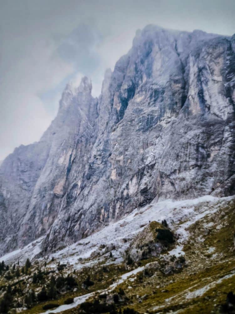 Steile, zerklüftete Bergfelsen der Dolomitenklassiker erheben sich über einen grasbewachsenen, felsigen Hang mit vereinzelten kleinen Bäumen unter einem wolkenverhangenen Himmel.