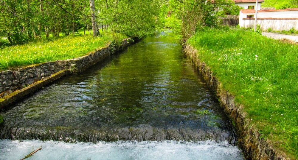 Im Kleinen Lautertal fließt ein klarer Bach zwischen grasbewachsenen Ufern und Steinmauern, umgeben von üppig grünen Bäumen und Pflanzen, mit einem rotgedeckten Haus und einem Laternenpfahl im Hintergrund - perfekt für eine ruhige Wanderung oder Auszeit.