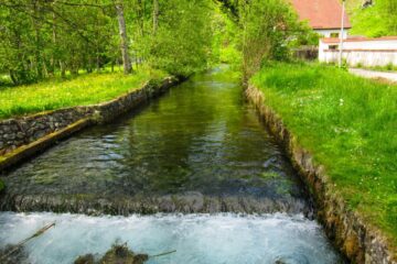 Im Kleinen Lautertal fließt ein klarer Bach zwischen grasbewachsenen Ufern und Steinmauern, umgeben von üppig grünen Bäumen und Pflanzen, mit einem rotgedeckten Haus und einem Laternenpfahl im Hintergrund - perfekt für eine ruhige Wanderung oder Auszeit.