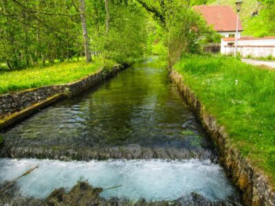 Im Kleinen Lautertal fließt ein klarer Bach zwischen grasbewachsenen Ufern und Steinmauern, umgeben von üppig grünen Bäumen und Pflanzen, mit einem rotgedeckten Haus und einem Laternenpfahl im Hintergrund - perfekt für eine ruhige Wanderung oder Auszeit.