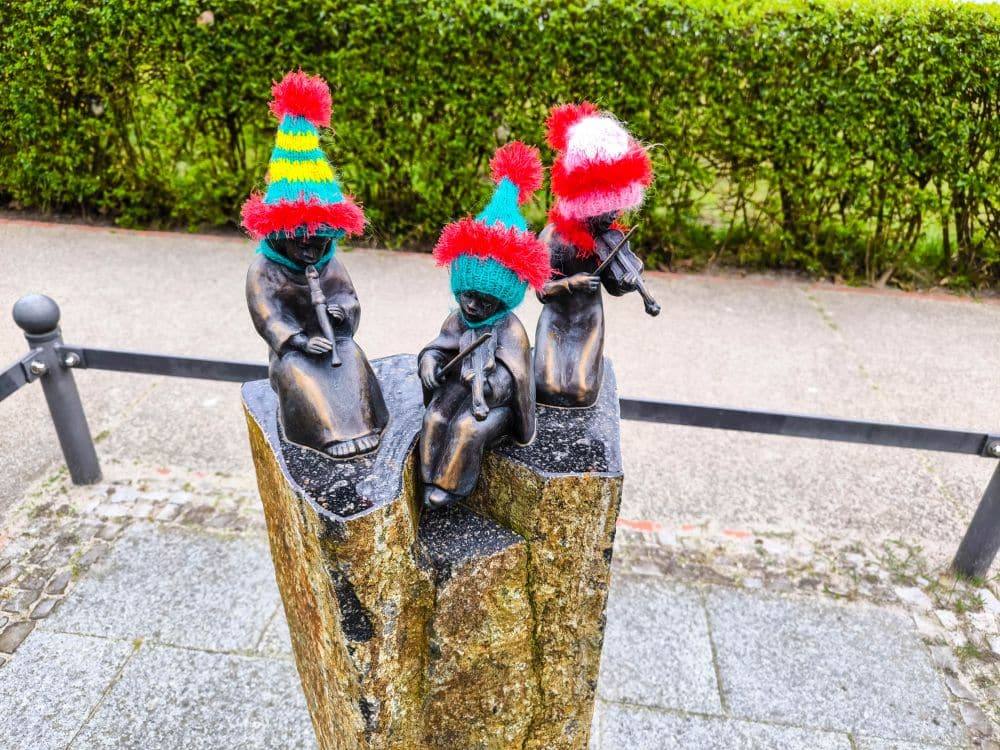 Auf einem Steinsockel in Binz stehen drei kleine Kinderstatuen aus Bronze, die jeweils eine bunte Strickmütze mit Bommeln tragen. Eine Statue spielt Geige. Im Hintergrund sind grüne Hecken und ein gepflasterter Weg zu sehen.