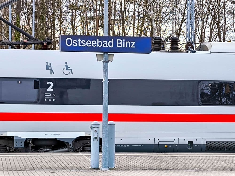 Ein Zug hält an einem Bahnsteig in Binz, auf dem ein blaues Schild mit der Aufschrift Ostseebad Binz steht. An der Seite des Zuges sind Symbole für Barrierefreiheit und zweite Klasse angebracht, während im Hintergrund Bäume stehen.