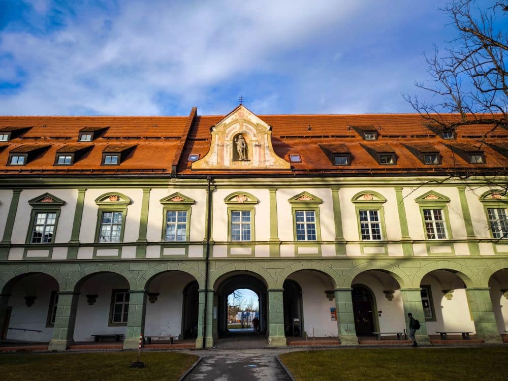 Ein historisches Barockgebäude mit rotem Ziegeldach, Rundbogenfenstern und einem zentralen Torbogeneingang. Die Fassade des Klosters Benediktbeuern hat grüne Akzente, eine kleine Statue über dem Eingang und Berge, die sich unter blauem Himmel und Wolken erheben.