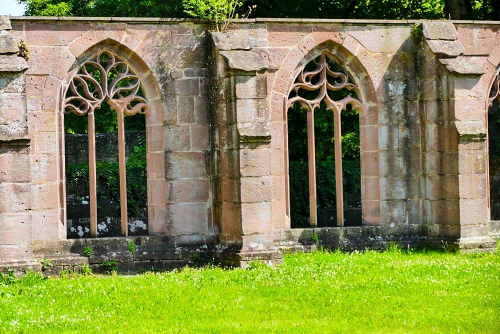 Auf einer Wiese in Hirsau, umgeben von Grün und Bäumen, stehen zwei große, gewölbte steinerne Fensterrahmen mit verschlungenen Mustern, die Teil einer alten, verwitterten Hauswand sind.