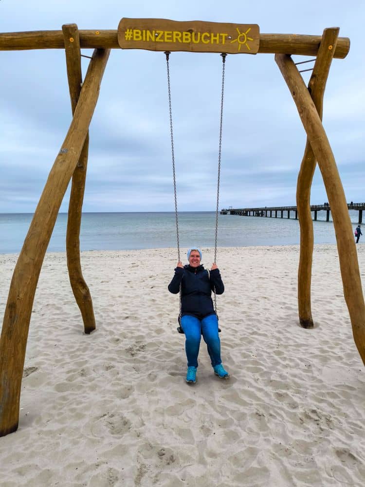 Eine Person sitzt auf einer großen Holzschaukel am Sandstrand von Binz, mit ruhigem Meer und Seebrücke im Hintergrund. Die Schaukel hat ein gelbes #BINZERBUCHT-Schild an der Spitze. Das Wetter ist bewölkt und kühl.