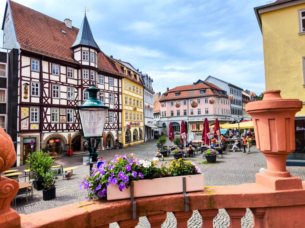 Ein bezaubernder europäischer Stadtplatz in Fulda mit farbenfrohen, historischen Fachwerkhäusern, Cafés im Freien, die von Sonnenschirmen beschattet werden, und einem Pflanzgefäß mit lila und weißen Blumen auf einem Steingeländer im Vordergrund.