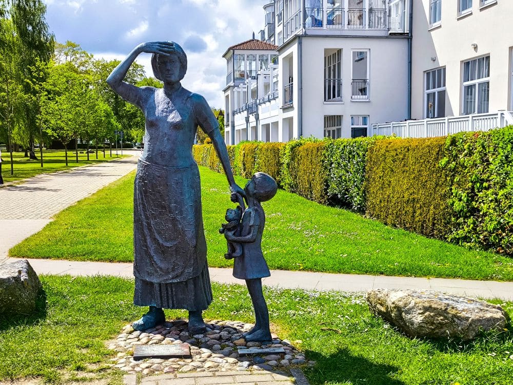 Eine Bronzestatue in Kühlungsborn zeigt eine Frau, die ihre Augen beschattet, und ein Kind, das einen Teddybär hält. Sie stehen auf einem Gehweg mit Gras, Sträuchern und der Ostseeküste sowie einem weißen Gebäude im Hintergrund.