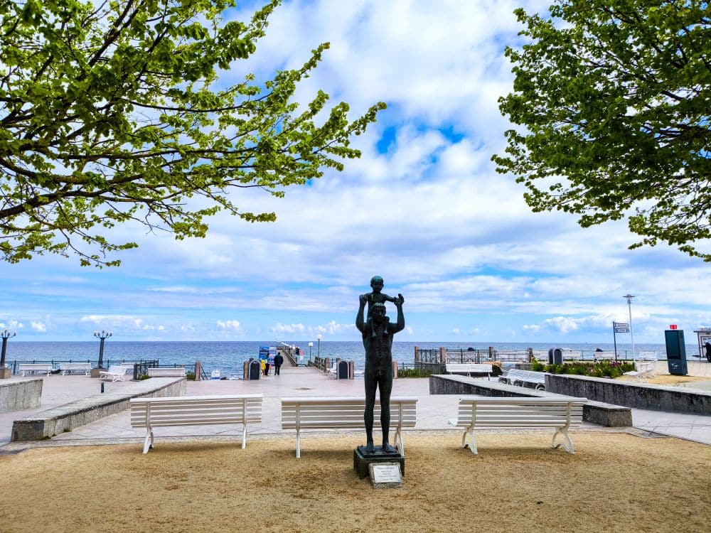 Eine Statue, die ein Kind auf den Schultern trägt, steht zwischen zwei Bänken auf einem Sandweg in Kühlungsborn, mit Blick auf eine Seebrücke, die sich unter einem teilweise bewölkten Himmel bis zur Ostsee erstreckt, eingerahmt von grünen Bäumen - perfekt für eine Auszeit.