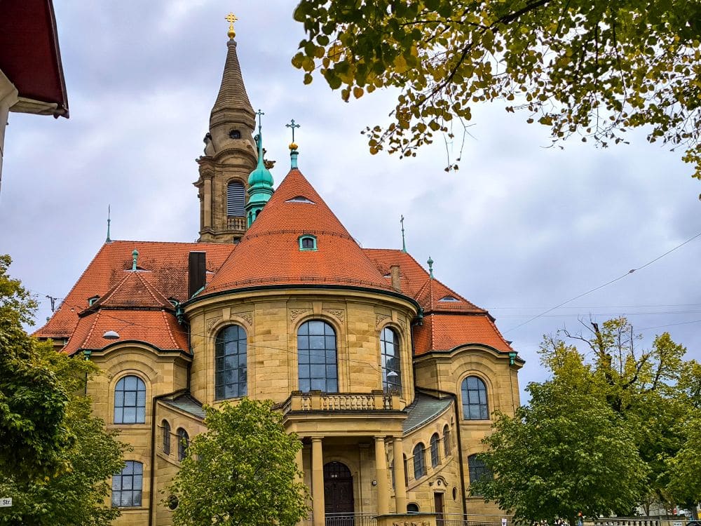 Eine historische Kirche mit rotem Ziegeldach, gewölbten Fenstern und einem hohen Glockenturm steht inmitten grüner Bäume und erinnert an den Barockcharme der berühmten Ludwigsburger Gärten unter einem bewölkten Himmel.