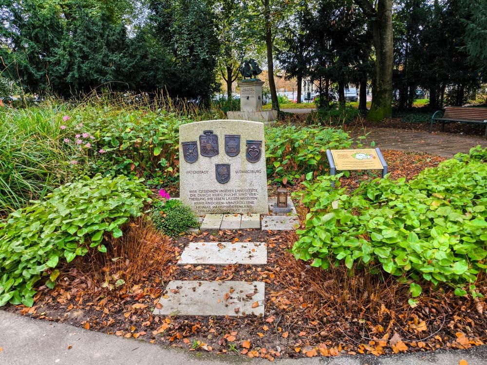 Ein steinernes Kriegerdenkmal mit militärischen Wappen und Inschriften steht in einem Park in Ludwigsburg, umgeben von grünen Büschen, heruntergefallenen Blättern und Informationstafeln. Im Hintergrund sind Bäume, Gärten und eine Bank zu sehen.
