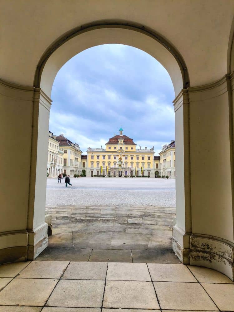 Blick durch einen großen Torbogen auf das große, gelbe Gebäude des Barock Ludwigsburg mit grüner Kuppel, das in einem weitläufigen Hof mit Kopfsteinpflaster unter einem bewölkten Himmel liegt. Ein paar Leute schlendern in Richtung der Gärten in der Ferne.