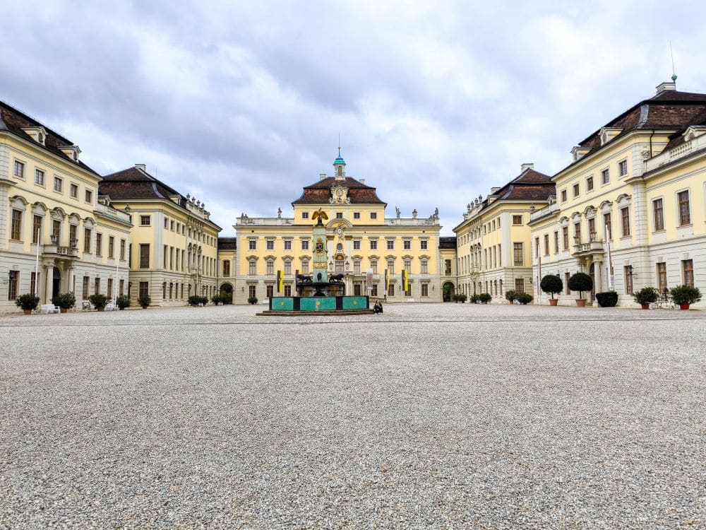 Ein großes Barockschloss in Ludwigsburg mit gelben und weißen Wänden, einem zentralen Uhrenturm und symmetrischen Flügeln. Ein Springbrunnen ziert den geräumigen Kieshof unter einem wolkenverhangenen Himmel.