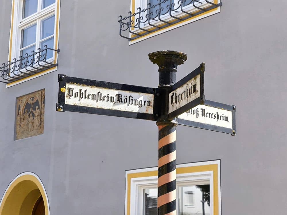 Ein schwarz-weiß gestreifter Wegweiser mit drei verblassten Richtungsschildern steht vor einem grauen Gebäude mit weiß umrandeten Fenstern und dekorativen Akzenten, die an den historischen Charme der Klosterstadt Neresheim erinnern.