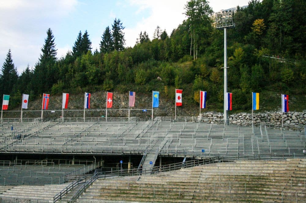 Leere Tribünenreihen in einem Freiluftstadion in Oberstdorf, mit mehreren Nationalflaggen über den Sitzen. Hinter den Fahnen vervollständigen ein grüner Alpenhügel und ein hoher Lichtmast die malerische Kulisse.