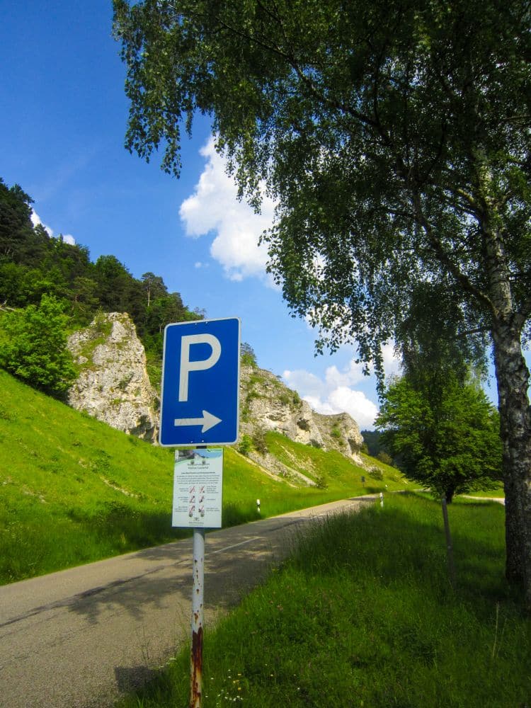 Ein blaues Parkplatzschild mit Rechtspfeil steht an einer Landstraße im Kleinen Lautertal, umgeben von grünen Wiesen, Bäumen und felsigen Hügeln - ein einladender Ort für eine Wanderung oder eine entspannende Auszeit unter dem strahlend blauen Himmel.