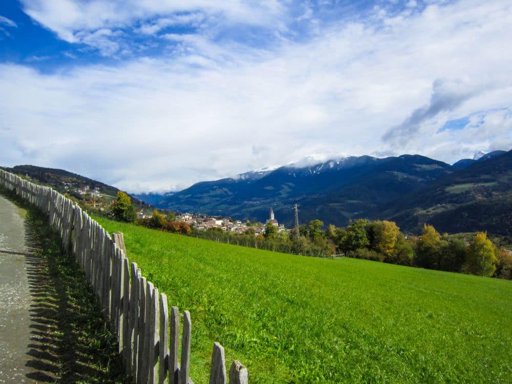 Eine sattgrüne, von einem Holzzaun gesäumte Wiese erstreckt sich in Richtung Feldthurns, einem kleinen Ort am Fuße der baumbestandenen Berge am malerischen Keschtnweg in Südtirol, und das alles unter einem teilweise bewölkten Himmel.
