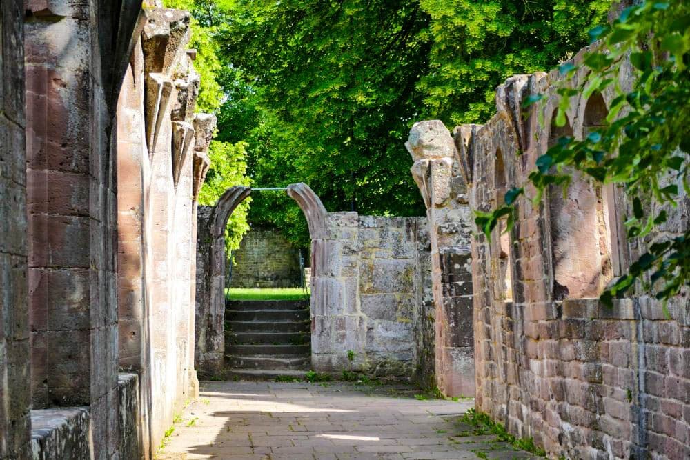 Alte steinerne Ruinen von Hirsau mit gewölbten Toren und verwitterten Mauern, umrahmt von grünen Bäumen und in Sonnenlicht getaucht. Steintreppen führen durch einen Bogen hinauf auf eine dahinter liegende Wiese.