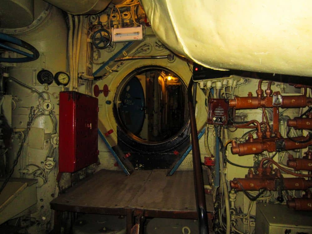 Innenansicht eines U-Boots aus der Peenemünder Zeit mit einer runden Luke, Rohren, Messgeräten, Ventilen und elektrischen Schalttafeln an beigen Wänden, mit einem roten Kasten auf der linken Seite und einem schmalen Gang, der durch die Luke führt.