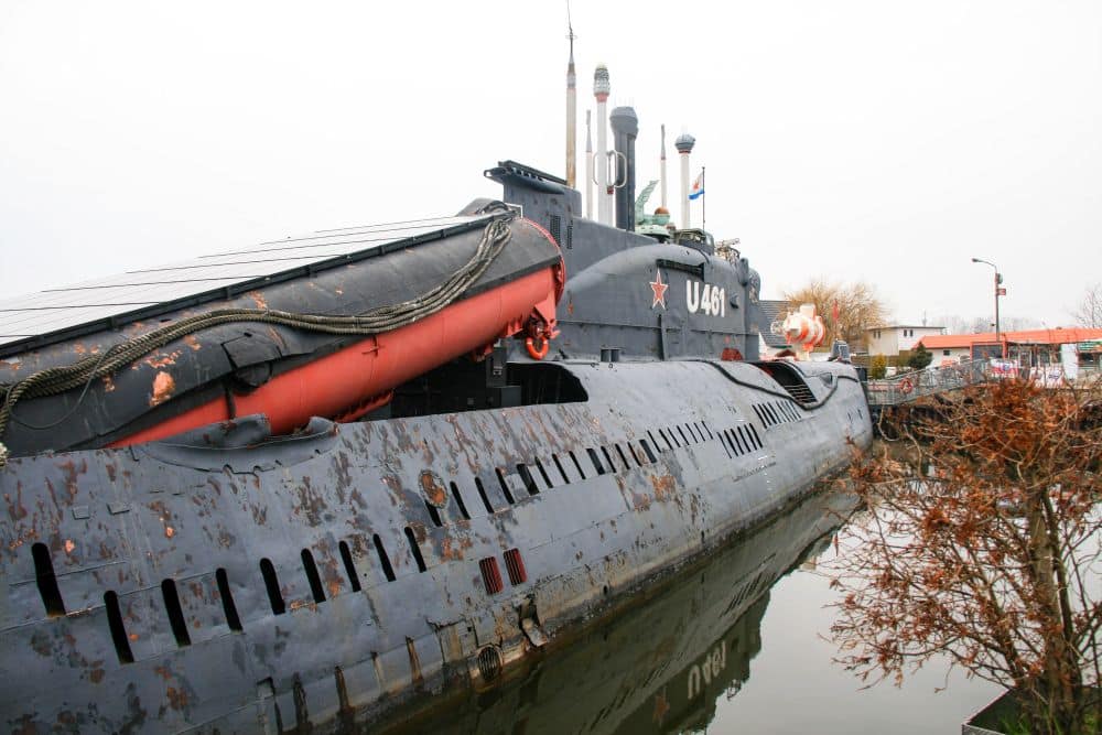 Ein großes, verwittertes U-Boot mit der Bezeichnung U461 liegt in Peenemünde im Wasser. Auf seinem Deck sind rote Rettungsboote montiert, und der Kommandoturm ist mit Antennen und Periskopen bestückt. In der Nähe sind spärliche Bäume und Gebäude zu sehen.