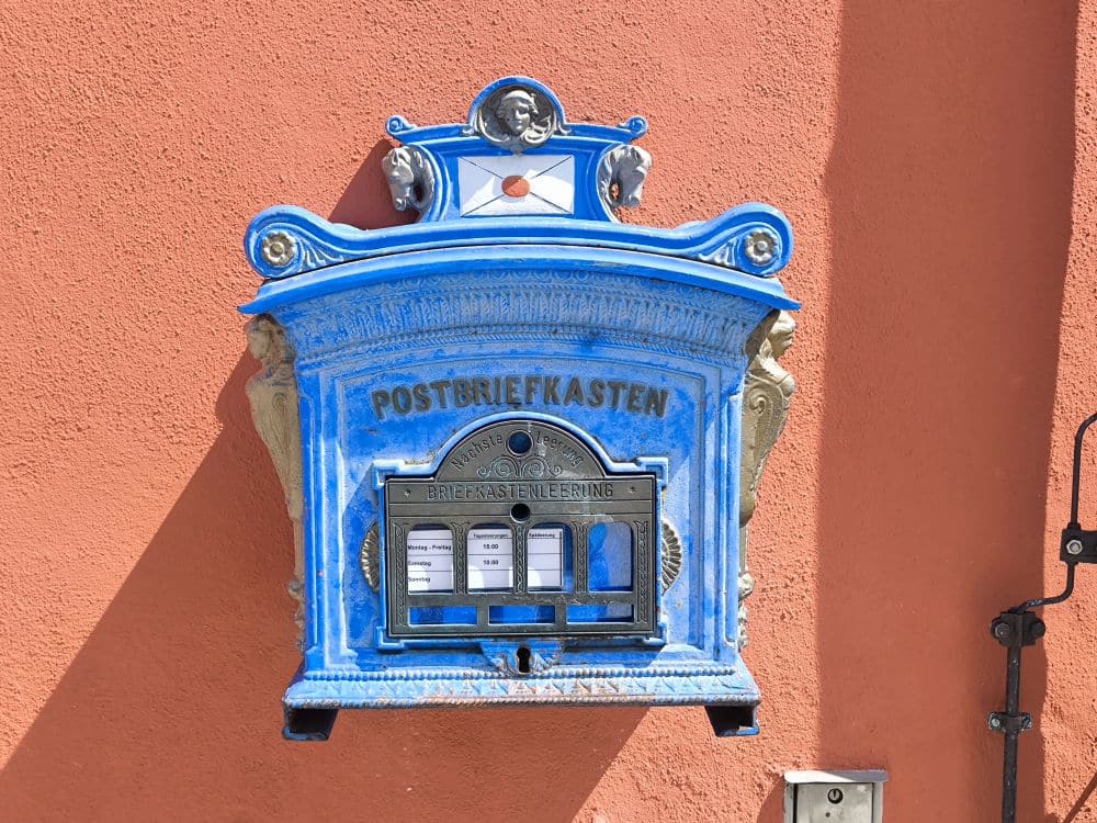 Ein alter blauer deutscher Briefkasten mit der Aufschrift POSTBRIEFKASTEN ist an einer orangefarbenen Wand in Nördlingen angebracht. Er ist mit dekorativen Elementen und drei beschrifteten Schlitzen für die Post versehen.