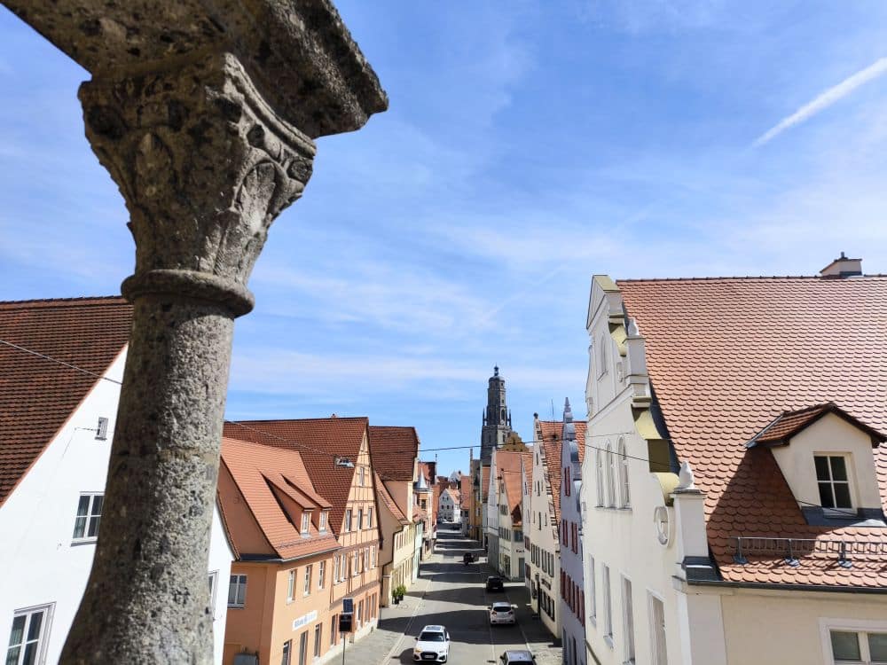Blick auf eine enge Straße in Nördlingen, die von bunten Häusern mit roten Ziegeldächern gesäumt ist, von einer erhöhten Position neben einer Steinsäule aus gesehen, unter einem klaren blauen Himmel. In der Ferne erhebt sich ein Kirchturm.