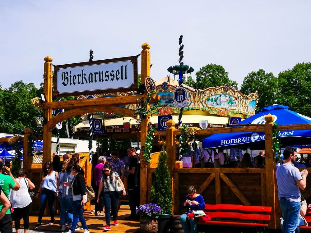 Menschenmassen versammeln sich auf einem Oktoberfest im Freien mit einem großen Holzschild mit der Aufschrift Bierkarussell, einem geschmückten Karussell im Hintergrund und blauen Hofbräu München-Regenschirmen, die belebte Sitzbereiche beschatten.