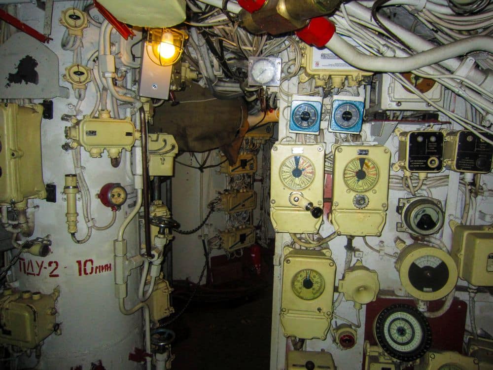 Das Bild zeigt das Innere eines U-Boots aus der Peenemünde-Ära, voll mit verschiedenen Ziffernblättern, Messgeräten, Knöpfen und Rohren an den Wänden, alles auf engstem Raum und beleuchtet von einer einzigen kleinen gelben Lampe.