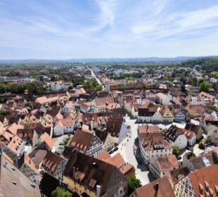 Luftaufnahme einer europäischen Stadt mit roten Dächern und engen Straßen - genießen Sie den Ausblick von Daniel in Nördlingen, umgeben von grünen Bäumen und fernen Hügeln unter einem blauen Himmel mit Wolkenfetzen.