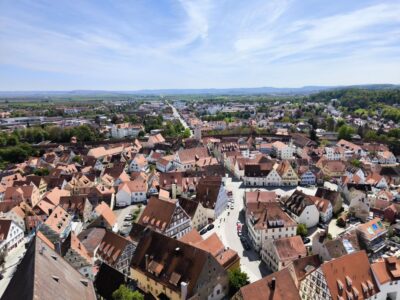 Luftaufnahme einer europäischen Stadt mit roten Dächern und engen Straßen - genießen Sie den Ausblick von Daniel in Nördlingen, umgeben von grünen Bäumen und fernen Hügeln unter einem blauen Himmel mit Wolkenfetzen.