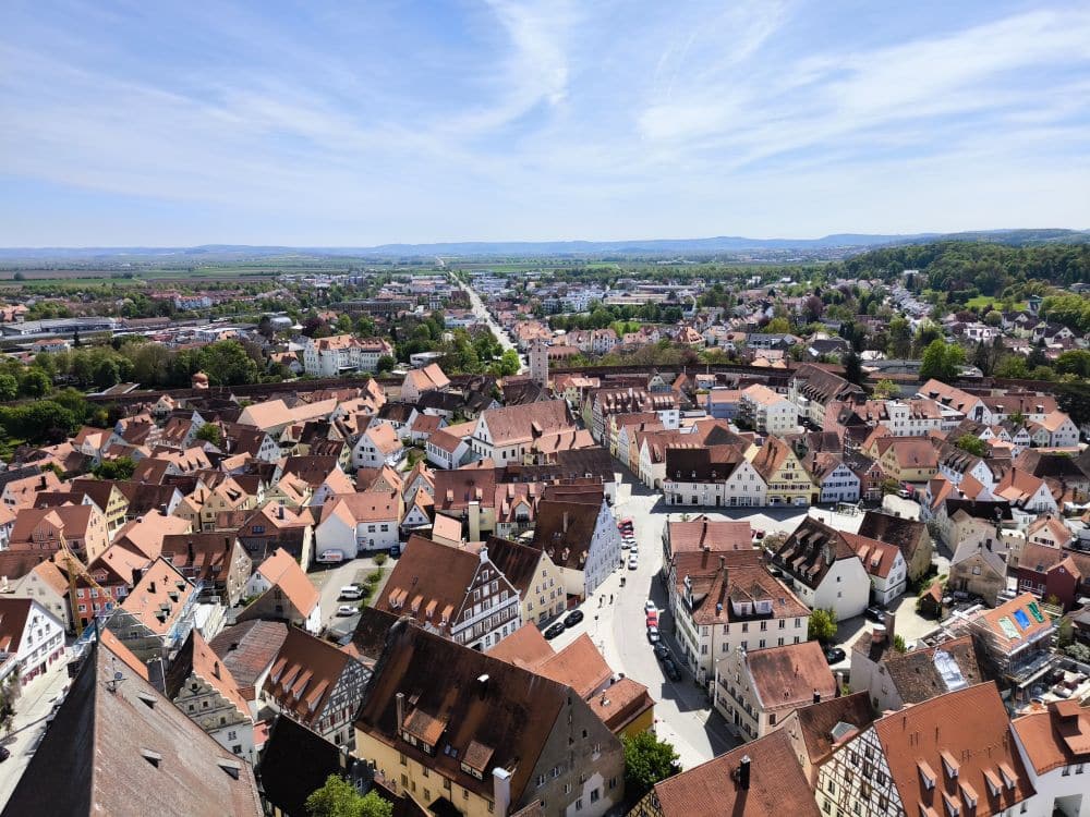 Luftaufnahme einer europäischen Stadt mit roten Dächern und engen Straßen - genießen Sie den Ausblick von Daniel in Nördlingen, umgeben von grünen Bäumen und fernen Hügeln unter einem blauen Himmel mit Wolkenfetzen.
