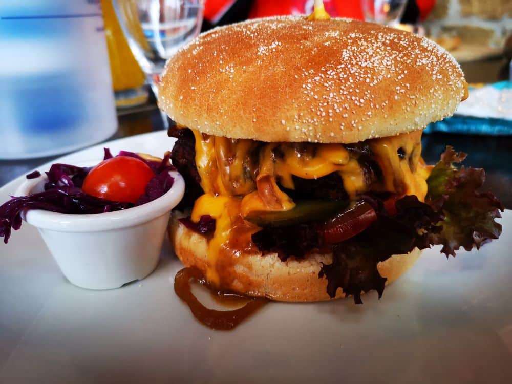 Ein Cheeseburger mit geschmolzenem Cheddar, Salat, Essiggurken und Zwiebeln im Brötchen, serviert mit Krautsalat und einer Kirschtomate - perfekt, um ihn in einem Ihrer Lieblingsrestaurants auf der Insel Rügen zu genießen.