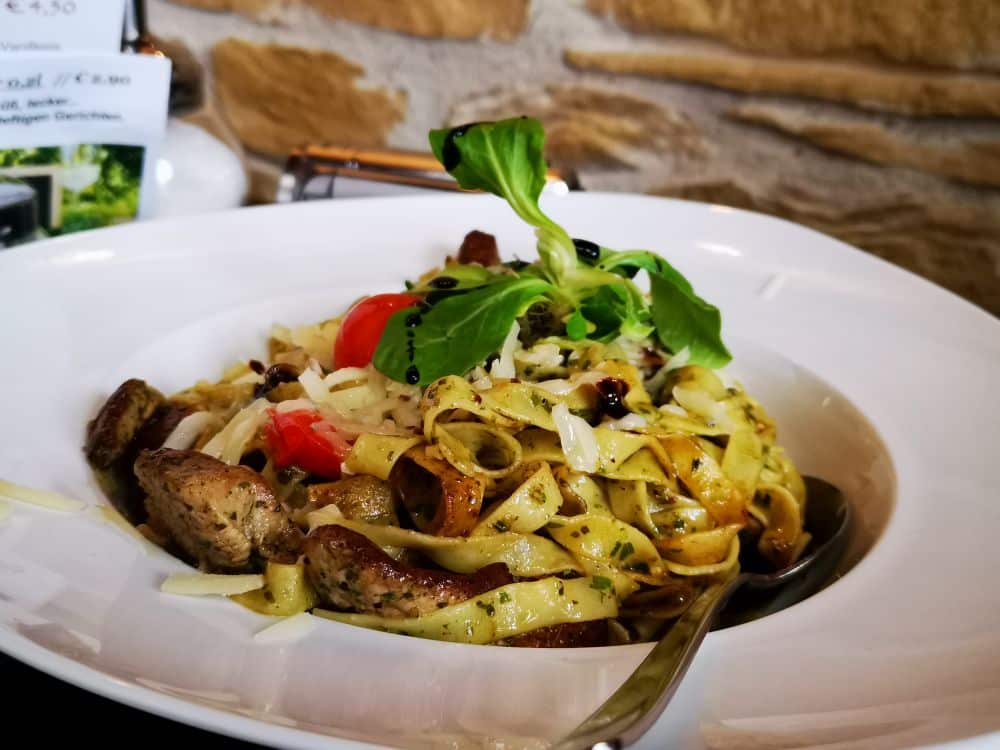 Eine weiße Schale gefüllt mit Tagliatelle-Nudeln, gegrilltem Fleisch, Kirschtomaten, Käse und Grünzeug, garniert mit Balsamico-Glasur und einem Löffel darin - die klassische Küche der Spitzenrestaurants auf der schönen Insel Rügen. Steinmauer im Hintergrund.